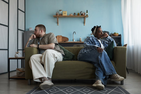 Caucasian young adult man and Black young adult woman sitting on sofa facing away from each other with arms crossed, both looking upset and avoiding eye contact during conflictの写真素材