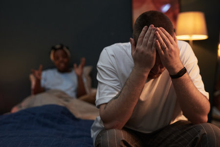 Caucasian young adult man sitting on bed covering face with hands, showing distress while Black young adult woman sitting behind gesturing with hands during argument in bedroomの写真素材