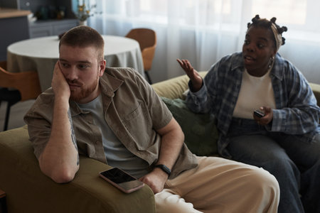 Caucasian young adult man sitting on sofa looking away with hand on face, while Black young adult woman gesturing and talking beside him during conflict in living roomの写真素材
