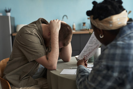 Caucasian young adult man sitting at table holding head in hands while Black young adult woman sitting across pointing at document, both appearing engaged in intense discussionの写真素材