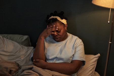 Black woman sitting on bed covering face with hand appearing distressed, showing signs of emotional conflict, wearing headband, illuminated by lamp, suggesting relationship struggleの写真素材
