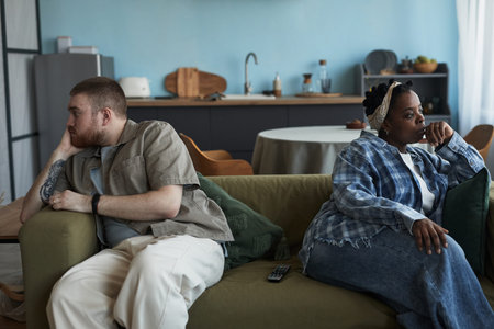 Caucasian young adult man and Black young adult woman sitting on sofa facing away from each other with arms crossed and serious expressions, showing conflict in relationshipの写真素材