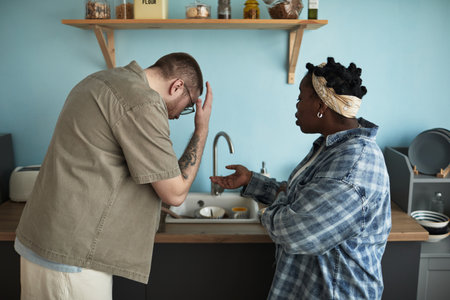 Middle aged Caucasian man holding head in frustration while middle aged Black woman gesturing with hand during argument in kitchen, both standing near sink, expressing conflictの写真素材