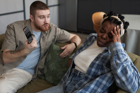 Middle aged Caucasian man holding remote control gesturing while middle aged Black woman sitting beside him touching forehead appearing upset during conflict on couchの写真素材