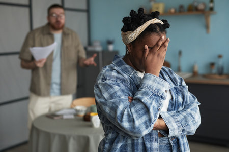 Black young adult woman standing with arms crossed and hand covering face, Caucasian young adult man in background gesturing and holding papers, both appearing to be in conflictの写真素材