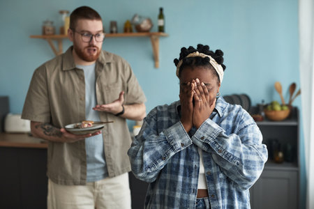 Caucasian young adult man gesturing while holding plate, Black young adult woman standing in foreground covering face with hands, both appearing upset during conflict in kitchenの写真素材