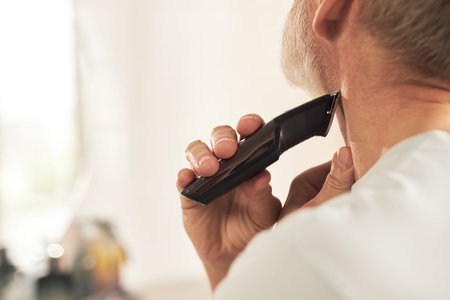Middle aged Caucasian man grooming neck with electric trimmer during skincare routine, hand holding...