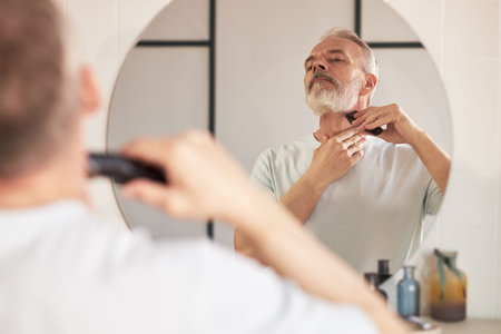 Senior Caucasian man grooming beard with electric trimmer in bathroom, standing in front of mirror and carefully shaping facial hair as part of daily beauty and skincare routineの写真素材