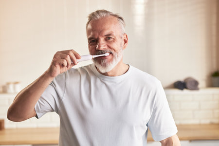 Portrait of senior Caucasian man smiling while brushing teeth with electric toothbrush in modern bathroom, demonstrating daily beauty and skincare routine for oral hygieneの写真素材