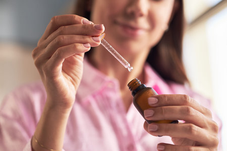 Young adult Caucasian woman holding glass dropper above skincare serum bottle, preparing to apply...