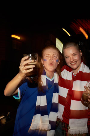 Two young Caucasian women celebrating together, one raising glass and making expressive face, both wearing soccer scarves and face paint, standing close in festive environment in sports pubの写真素材
