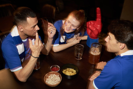 Three young soccer fans including two Caucasian men and one woman sitting at table in pub, talking...