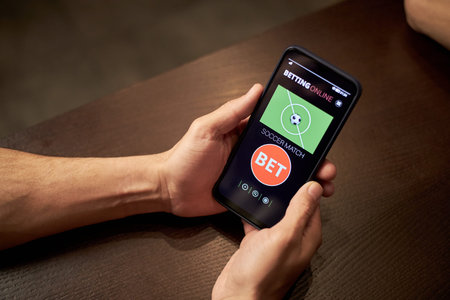 Hands of anonymous man holding smartphone displaying online soccer betting application, sitting at table in bar, engaging in sports gambling activityの写真素材