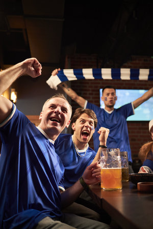 Group of Caucasian and multiethnic young men celebrating goal in sports bar, raising fists and cheering while watching soccer match, one man holding scarf, beer glasses on tableの写真素材