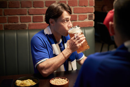 Caucasian young adult man, wearing blue soccer fan scarf, drinking beer from large glass mug at table with snacks in sports bar, sitting with friend during match viewingの写真素材