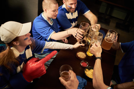 Group of multiethnic young men and women wearing soccer fan gear celebrating together, raising beer...
