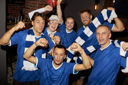 Group portrait of multiethnic sports fans celebrating, wearing blue soccer jerseys and scarves, raising fists and smiling, showing excitement while supporting their team in barの写真素材