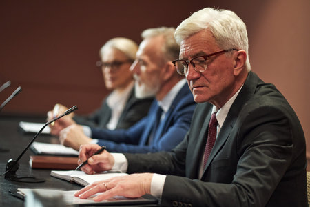 Senior Caucasian man sitting at conference table, writing notes during political meeting with two...