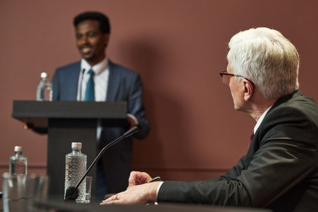 Middle aged Black man standing at podium speaking, while middle aged Caucasian man sitting at table...