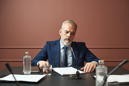 Middle aged Caucasian man sitting at table reading documents during political meeting, microphones and water bottles on desk, wearing suit and tie, focused expressionの写真素材