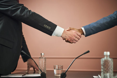Middle aged Caucasian man and middle aged Black man shaking hands over conference table, microphones...