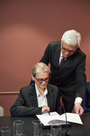 Middle aged Caucasian woman sitting at table signing document while senior Caucasian man standing beside her pointing at paper, both wearing business suits in formal meeting settingの写真素材