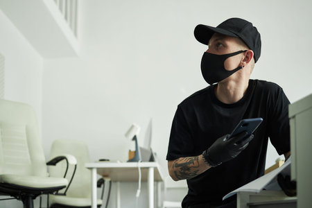 Male intruder wearing black mask and gloves holding smartphone, taking photos of corporate...