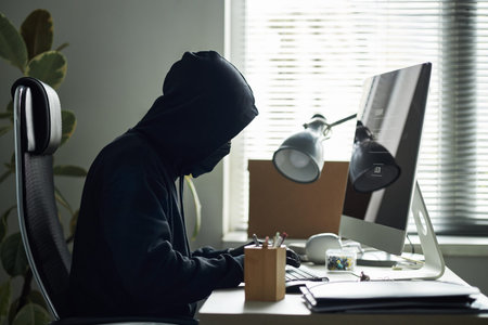 Young man wearing black hooded sweatshirt, obscuring face, sitting at desk, hacking into desktop...