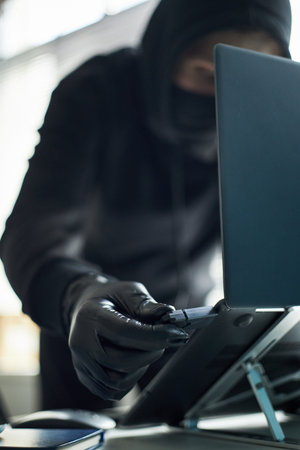 Unrecognizable man wearing black gloves and hoodie inserting USB flash drive with malware into laptop to hack corporate computer system, engaging in covert criminal activity at workspaceの写真素材