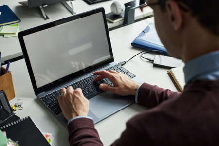 Unrecognizable young man working on laptop at desk, hands typing on keyboard, office supplies and...