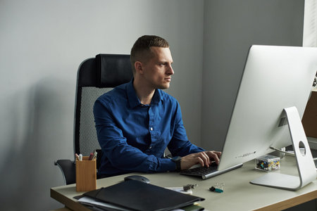 Caucasian young man sitting at desk working on computer, focusing on screen with hands on keyboard,...