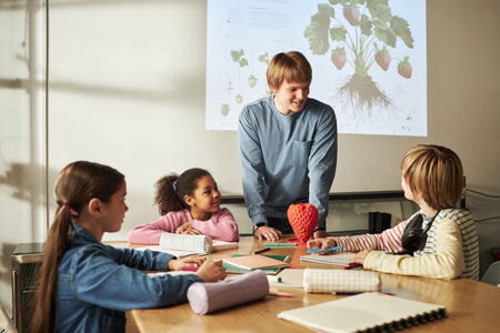 Caucasian young adult man teaching diverse group of children in classroom, students sitting at table...
