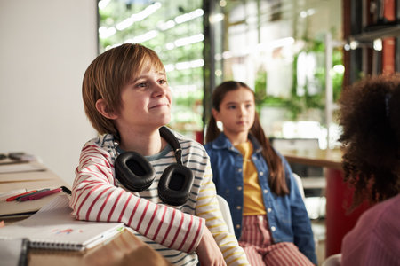 Caucasian boy with headphones sitting at desk smiling and looking to side, multiethnic group of children studying together in modern classroom, notebooks and supplies on tableの写真素材