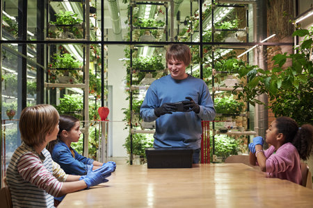 Young man standing and gesturing, while teaching group of children, wearing gloves at table in...