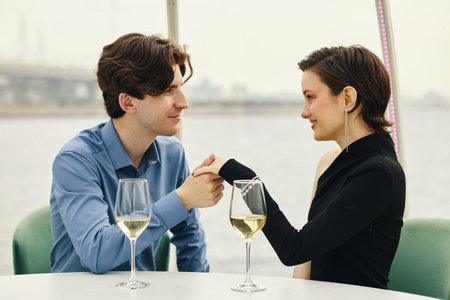 Young Caucasian man holding hand of young Caucasian woman while sitting at table with wine glasses, both smiling and making eye contact in outdoor waterfront settingの写真素材