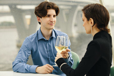 Caucasian young man and Caucasian young woman sitting at table clinking wine glasses, man smiling...