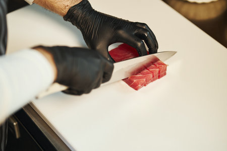 Caucasian man slicing raw fish with sharp knife on cutting board, wearing black gloves, preparing ingredients for meal, close up of hands and forearms in professional kitchenの写真素材