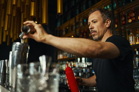 Middle aged Caucasian man preparing cocktail behind bar counter, holding shaker and bottle, focusing...