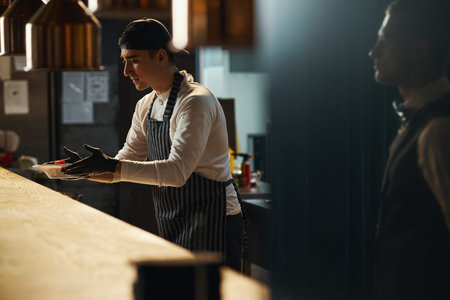 Young adult Caucasian man wearing apron and cap serving food at restaurant counter, extending hands...
