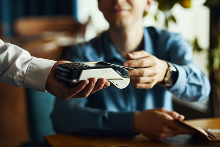Caucasian young adult man handing credit card to another holding payment terminal, making contactless transaction in indoor setting, focus on hands and deviceの写真素材