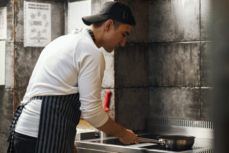 Young adult man wearing apron standing in professional kitchen cooking food in frying pan on stovetop, concentrating on preparing meal in restaurant settingの写真素材
