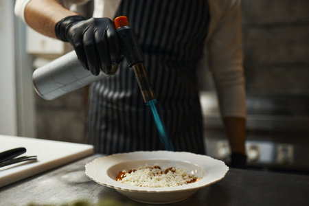 Caucasian young adult man wearing striped apron using culinary torch to caramelize dish in restaurant kitchen, right hand holding torch over plate, black gloves visibleの写真素材