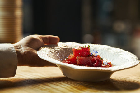 Caucasian male hand holding textured plate with gourmet fruit salad featuring watermelon cubes, raspberries, edible flowers, herbs, wooden table surface visibleの写真素材