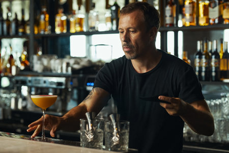Middle aged Caucasian man preparing cocktail behind bar counter, holding black saucer and arranging glassware, liquor bottles displayed on shelves in backgroundの写真素材
