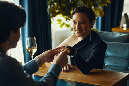Caucasian young adult woman smiling while holding hands with Caucasian young adult man across table in restaurant, wine glass on table, woman making eye contact with manの写真素材