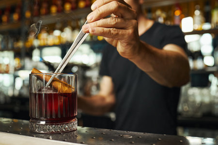 Caucasian young adult man stirring cocktail with cinnamon stick in glass using metal bar spoon,...