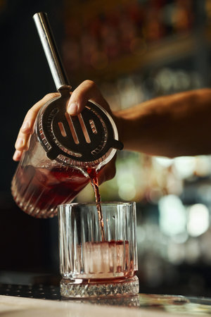 Caucasian young adult male hand pouring red cocktail from mixing glass through strainer into tumbler...