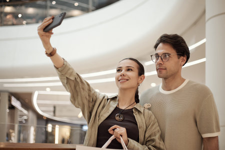 Young adult Caucasian woman smiling and holding smartphone taking selfie with young adult Caucasian man standing beside her in modern furniture store interiorの写真素材