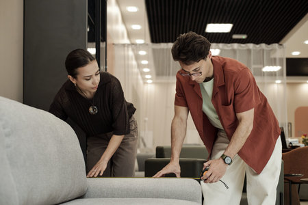 Caucasian man and Caucasian young adult woman measuring sofa together in furniture store, both...