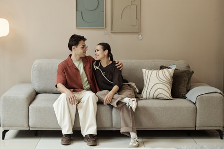 Young adult Caucasian man and woman sitting together on sofa smiling and looking at each other in furniture store, man putting arm around womanの写真素材